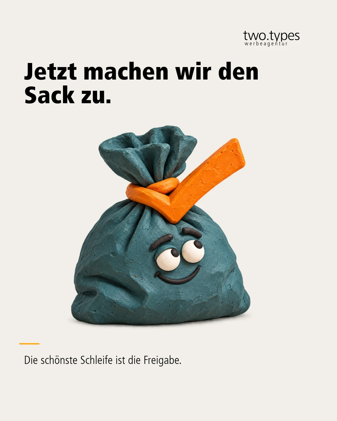 Ein petrolfarbener Sack mit Augen ist oben mit einer orangefarbenen Schleife in Häkchenform verschlossen.